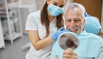 Man with white hair smiling into mirror with dentist next to him pointing to his teeth