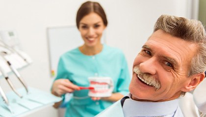 Man with gray hair smiling as dentist demonstrates how to brush dentures
